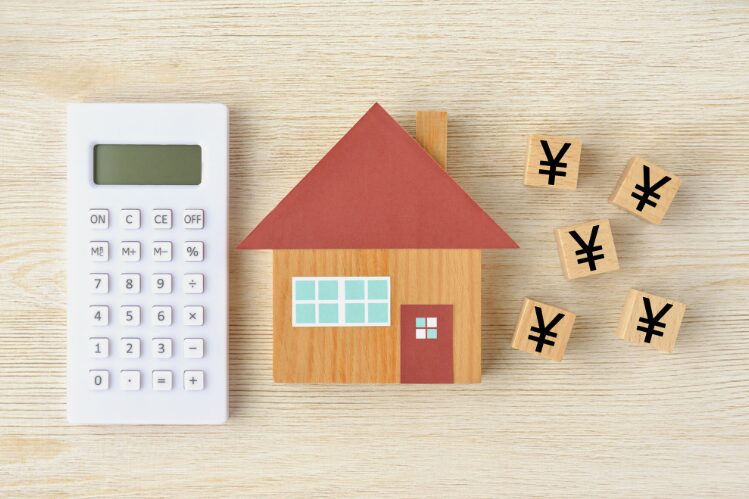 木製の家の模型と電卓、周囲に並べられた円マークの積み木が置かれている様子。住宅費用や資金計画をイメージした構図