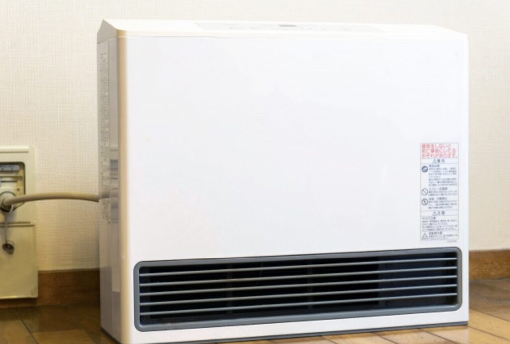 室内に設置された白いガスファンヒーターの写真。床近くのコンセントに接続され、吹き出し口が前面下部にある構造で、家庭用暖房機器として使用されている様子。